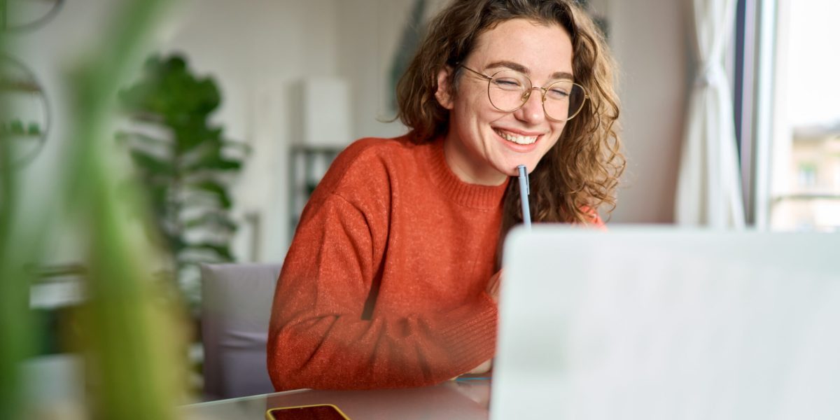 Happy young woman using laptop sitting at desk writing notes while watching webinar, studying online, looking at pc screen learning web classes or having virtual call meeting remote working from home.